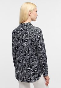 Chemise à manches longues noire avec un motif géométrique blanc, dotée d'un col et d'un ourlet arrondi. Tissu lisse, coupe décontractée, détail de l'empiècement au dos.