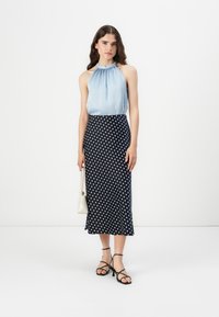 Lyseblå halterneck topp, marineblå polka-dot midi skjørt, svarte stroppesandaler og hvit håndveske; glatt stoff, tettsittende silhuett og volanghalset.