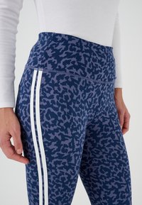Donna che indossa leggings blu con stampa leopardata e strisce laterali bianche, con una maglietta bianca a maniche lunghe, in piedi con le mani rilassate ai lati.