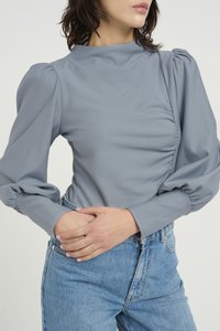 Light grey blouse with puffed sleeves and a mock neck; features gathered detail on the side. Worn with high-waisted blue denim jeans.