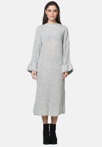 Langes, graues Strickkleid aus strukturiertem Stoff, mit weiten Ärmeln und geradem Saum. Das Model trägt hohe, schwarze Stiefel mit Absatz.