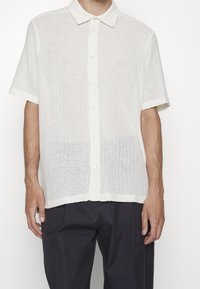 Camisa blanca ligera de botones, con un patrón texturizado de tejido abierto y mangas cortas, que presenta un cuello clásico y un dobladillo recto.