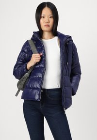 Marineblå puffjakke med hette, med en skinnende tekstur, quiltet design, frontglidelås og lommer til hender, båret over en hvit topp og jeans.