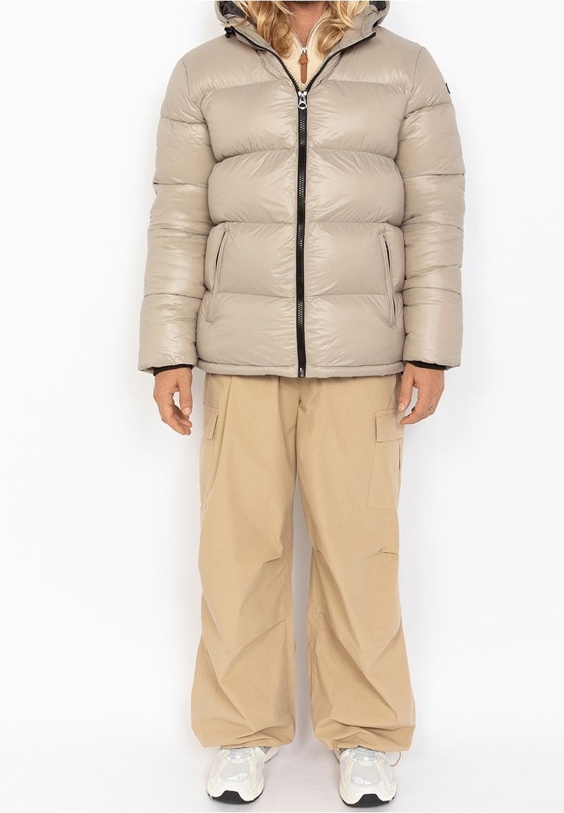 Personne portant une doudoune beige avec une fermeture éclair noire, un pantalon cargo beige et des baskets blanches, debout sur fond blanc.