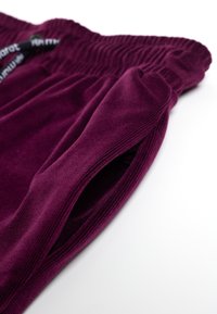 Bordeauxrode veloursbroek met elastische tailleband en zijzakken. Gladde textuur en geribbelde details langs de tailleband en randen.