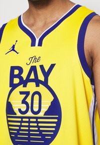 Maillot de basket-ball jaune avec un design en col en V, des accents bleus et un imprimé graphique avec "The BAY" et "30" en grandes lettres. Détail du logo Jordan.