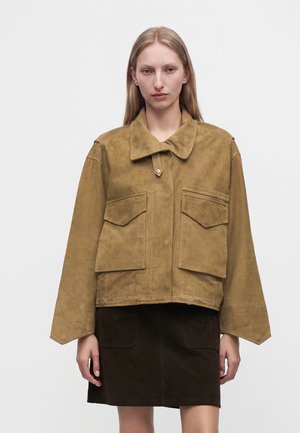 Frau mit glatten blonden Haaren, die eine braune Wildlederjacke mit großen Vorderentaschen und einen dunkelbraunen Rock trägt, stehend vor einem schlichten weißen Hintergrund.