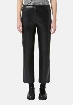 Personne portant un pantalon en cuir noir à jambes droites, des chaussettes noires et des chaussures noires à semelle épaisse, debout devant un fond uni.