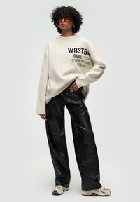 Överdimensionerad beige sweatshirt med svart grafisk text, kombinerad med löst sittande svarta byxor i imitationsläder och beiga sneakers med detaljer.