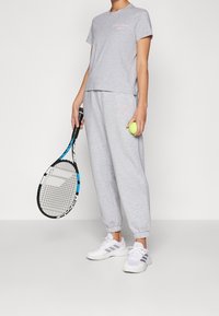 Person i grå sportswear, der holder en tennisketsjer i den ene hånd og en tennisbold i den anden, stående på en ensfarvet baggrund.