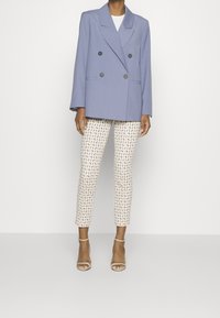 Lysblå double-breasted blazer med store sorte knapper, parret med mønstrede kremfarvede bukser med geometriske motiver og beige stroppede sandaler.