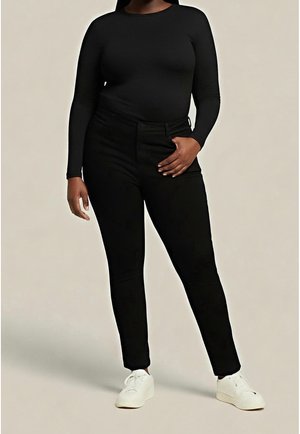 Frau trägt eng anliegendes schwarzes Langarmshirt, hoch taillierte schwarze Hose und weiße Turnschuhe, steht vor einfarbigem beigem Hintergrund.