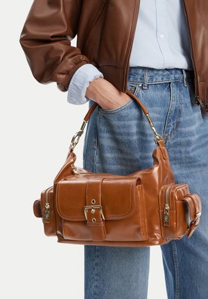 Woman wearing brown leather jacket and jeans holding a brown leather handbag with buckle and multiple zippered pockets.