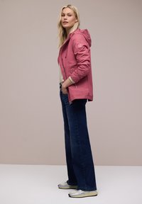 Junge Frau trägt eine rosa Kapuzenjacke, ein cremefarbenes Oberteil, dunkelblaue, weit geschnittene Jeans und silberne Turnschuhe, steht mit den Händen in den Jackentaschen.