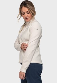 Weißer Fleece-Jacke mit hohem Kragen und Reißverschluss, mit dezenten Nähten und einem Logo auf der rechten Schulter, getragen mit dunklen Hosen.