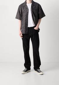 Korte mouwen zwart gemusterd shirt over een wit T-shirt, gecombineerd met zwarte jeans en zwarte sneakers. Het shirt heeft een subtiele luipaardprint.