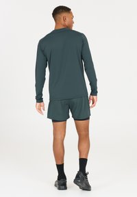 Dunkelgrünes Langarmhemd mit schwarzen Shorts. Synthetisches Material, figurbetontes Design. Schwarze Sportschuhe und Socken runden das Outfit ab.