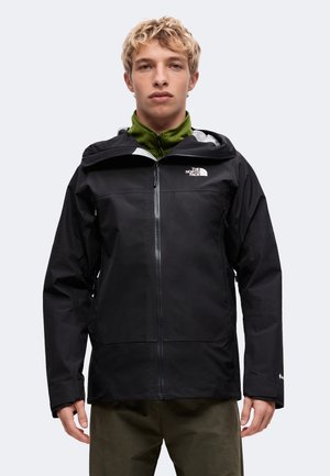 Młody mężczyzna ubrany w czarną wodoodporną kurtkę The North Face z kapturem i zielony polar z zamkiem pod spodem, stojący na jednolitym tle.