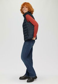 Granatowa kamizelka puchowa z wysokim kołnierzem, zestawiona z rdzawej koszuli z długim rękawem i niebieskimi dżinsami. Czarne buty mają teksturowany design.