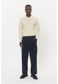 Beige polo tričko s dlhým rukávom a tromi gombíkmi, skombinované s tmavomodrými kordovými nohavicami a čiernymi šnurovacími topánkami, na bielom pozadí.