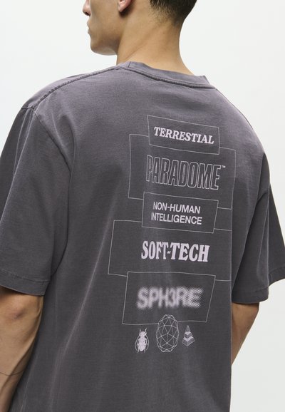 Moški z malce zbledelim črnim majico s praznimi belimi napisi "TERRESTRIAL," "PARADOME," "NON-HUMAN INTELLIGENCE," "SOFT-TECH," in "SPHERE" ter tremi geometrijskimi ikoni.