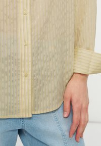 Ανοιχτό κίτρινο διαφανές πουκάμισο με κουμπιά και κάθετες ρίγες φορεμένο πάνω από ανοιχτό μπλε τζιν, με το χέρι χαλαρό στο πλάι.