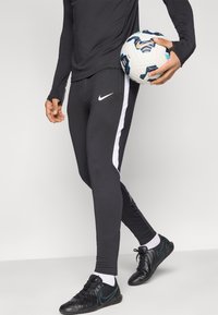 Μαύρο αθλητικό κολάν με λευκές ρίγες στο πλάι, σε συνδυασμό με μαύρα αθλητικά παπούτσια. Το άτομο κρατάει μια ποδοσφαιρική μπάλα κάτω από το χέρι του.