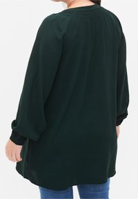 Tunika a maniche lunghe di colore verde scuro con una vestibilità ampia, tessuto testurizzato e scollo arricciato. Presenta polsini elastici e un orlo con spacco laterale.