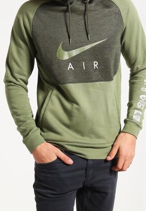 Personne portant un hoodie Nike Air vert olive avec un logo sur la poitrine et la manche, les mains dans les poches, assorti à un pantalon noir.