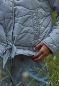 Lichtblauwe gewatteerde jas met een gestrikte taille, verticale stikdetails en een zachte textuur; gedragen over middelbleke denim jeans.