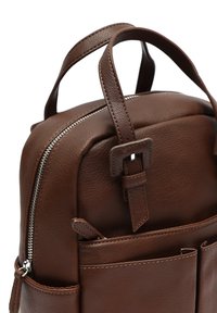 Sac à dos en cuir marron avec une surface texturée, compartiment principal zippé et deux poches avant. Il dispose de bretelles réglables et de poignées robustes.