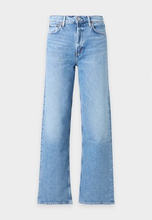 Jeune femme aux longs cheveux blonds portant un haut sans manches à col roulé rayé, un jean bleu à jambes larges et des chaussures marron pointues, debout les mains dans les poches.