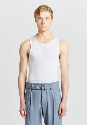 Jeune homme aux cheveux blonds courts porte un débardeur blanc et un pantalon bleu clair taille haute avec ceinture, debout sur un fond gris uni.
