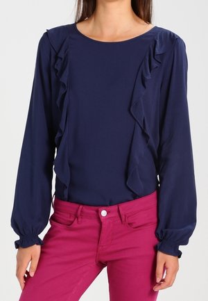 Femme portant un chemisier bleu marine à manches longues avec des volants sur le devant et un pantalon ajusté rose vif.