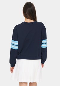 Felpa blu navy con strisce azzurre sulle maniche, tessuto in cotone, vestibilità rilassata, scollatura a girocollo, abbinata a una gonna bianca plissettata.
