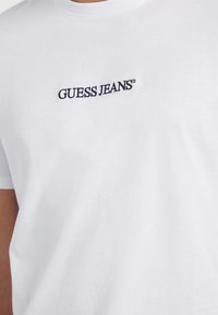 Balta kokvilnas T-kreklu ar apaļu kakla izgriezumu un īsām piedurknēm; uz krūtīm ir izšūts tumši zils "GUESS JEANS" logo.