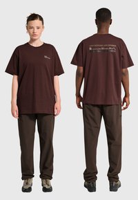 Två personer i mörkbruna vardagsbyxor och matchande oversized bruna T-shirts; text syns på framsidan och övre delen av ryggen på tröjorna.