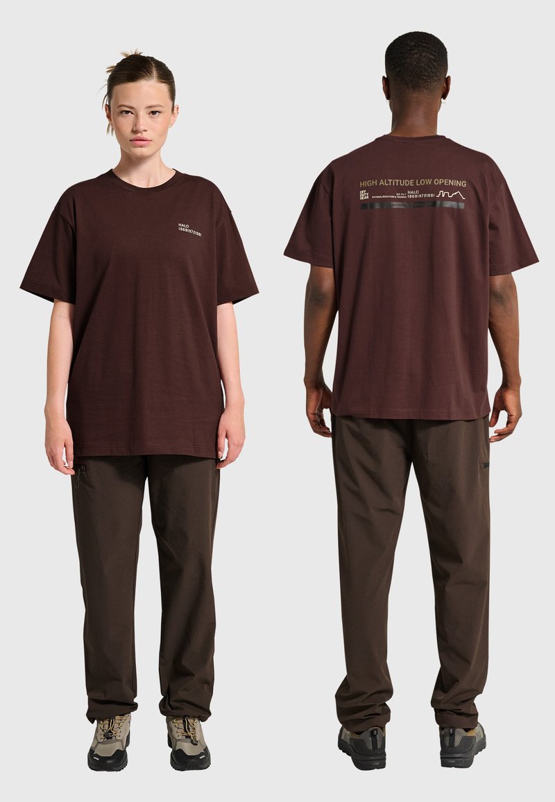 Två personer i mörkbruna vardagsbyxor och matchande oversized bruna T-shirts; text syns på framsidan och övre delen av ryggen på tröjorna.