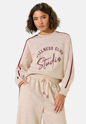 Frau mit lockigem Haar, die einen beigen Pullover und eine Hose mit maroonfarbenen Streifen trägt, auf dem Oberteil steht "Wellness Club Studio".