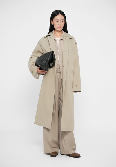 Μπεζ υπερμεγέθη παλτό, υφασμάτινη πλεκτή μπλούζα, παντελόνι φαρδιάς γραμμής, μαύρη δομημένη τσάντα στο αριστερό χέρι, καφέ επίπεδα παπούτσια, απλό φόντο.