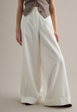 Frau trägt hochtaillierte, weit geschnittene weiße Hosen mit aufgekrempelten Säumen und eine braune, geknöpfte Weste, steht vor einem neutralen Hintergrund.