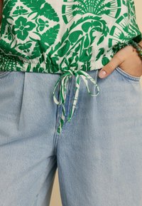 Green floral print top with cinched waist, paired with light blue high-waisted jeans. Notable drawstring detailing at the top.