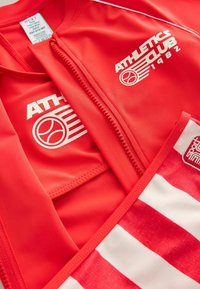 Punainen urheiluasu valkoisilla yksityiskohdilla. Varustettu vetoketjulla, "ATHLETICS CLUB" -logolla ja erottuvalla punaisella ja valkoisella raidallisella muotoilulla.