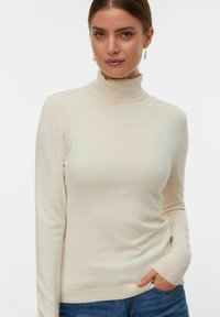 Suéter de cuello alto crema, hecho de una tela suave, con mangas largas y detalles acanalados en el cuello y los puños, combinado con jeans azules.