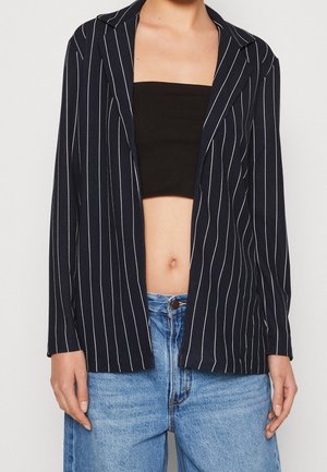 La mujer lleva un crop top negro, una americana abierta de raya diplomática azul oscuro y vaqueros de tiro alto azul claro, con las manos relajadas a los lados.