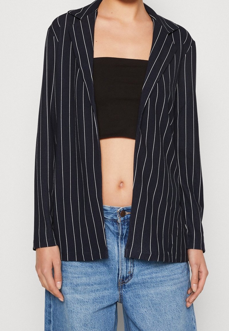 Femme portant un crop top noir, un blazer ouvert bleu marine à fines rayures, et un jean taille haute bleu clair, les mains détendues le long du corps.