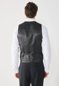 Gilet en satin noir à finition brillante, avec une boucle d'ajustement en argent, porté sur une chemise blanche, vu de dos.