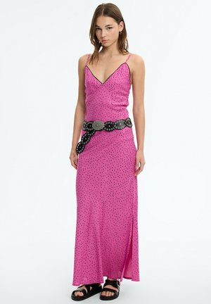Joven mujer con un vestido maxi rosa con lunares y tirantes finos, un cinturón negro con tachuelas y sandalias negras, posando sobre un fondo blanco.