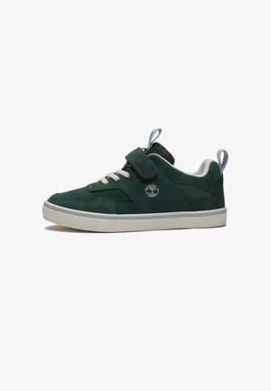 Zapatilla deportiva baja de color verde oscuro con suela blanca, cordones y una tira de velcro, con un logo de árbol en el lateral.