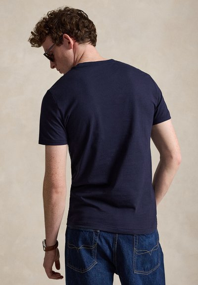 Mand med krøllet hår iført mørke solbriller, navy kortærmet t-shirt og blå jeans, stående med ryggen mod beskueren.